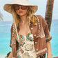 A woman in the Nevis One Piece by Hunter Bell NYC, paired with a wide-brimmed hat, large sunglasses, and a brown shell-embellished cover-up stands by the beach, framed by palm trees and the ocean.