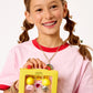A smiling girl with braces and pigtails holds a yellow Super Smalls box featuring the Paint it Heartfelt Mini Nail Kit, with glittery nail polish, heart-themed nail stickers, lip balms, and accessories, against a plain background.