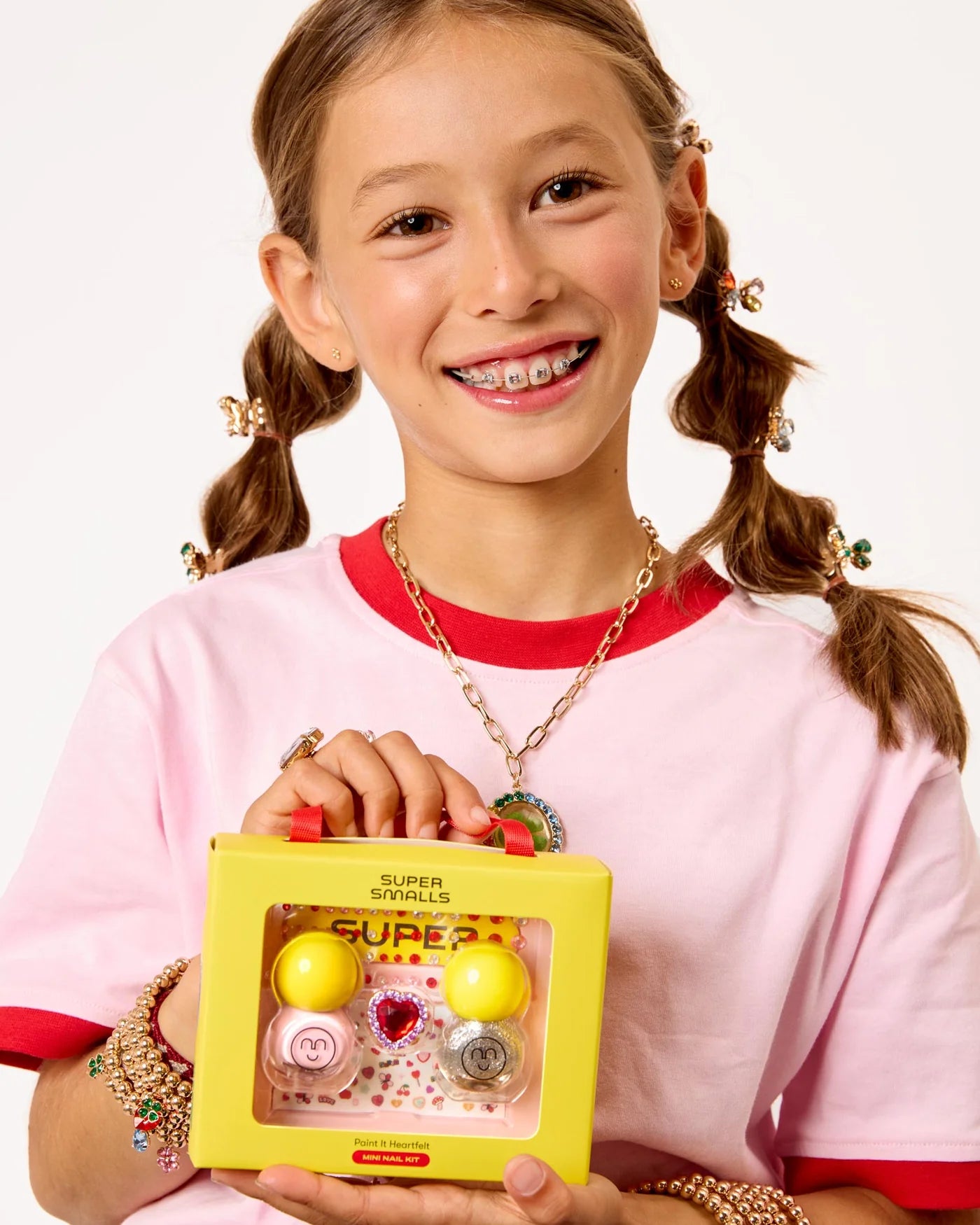 A smiling girl with braces and pigtails holds a yellow Super Smalls box featuring the Paint it Heartfelt Mini Nail Kit, with glittery nail polish, heart-themed nail stickers, lip balms, and accessories, against a plain background.