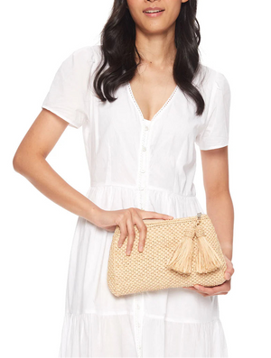 A woman in a short-sleeve white dress holds the Mar Y Sol Noa Clutch, featuring tassel accents—the ideal accessory for warm weather getaways—against a plain background.