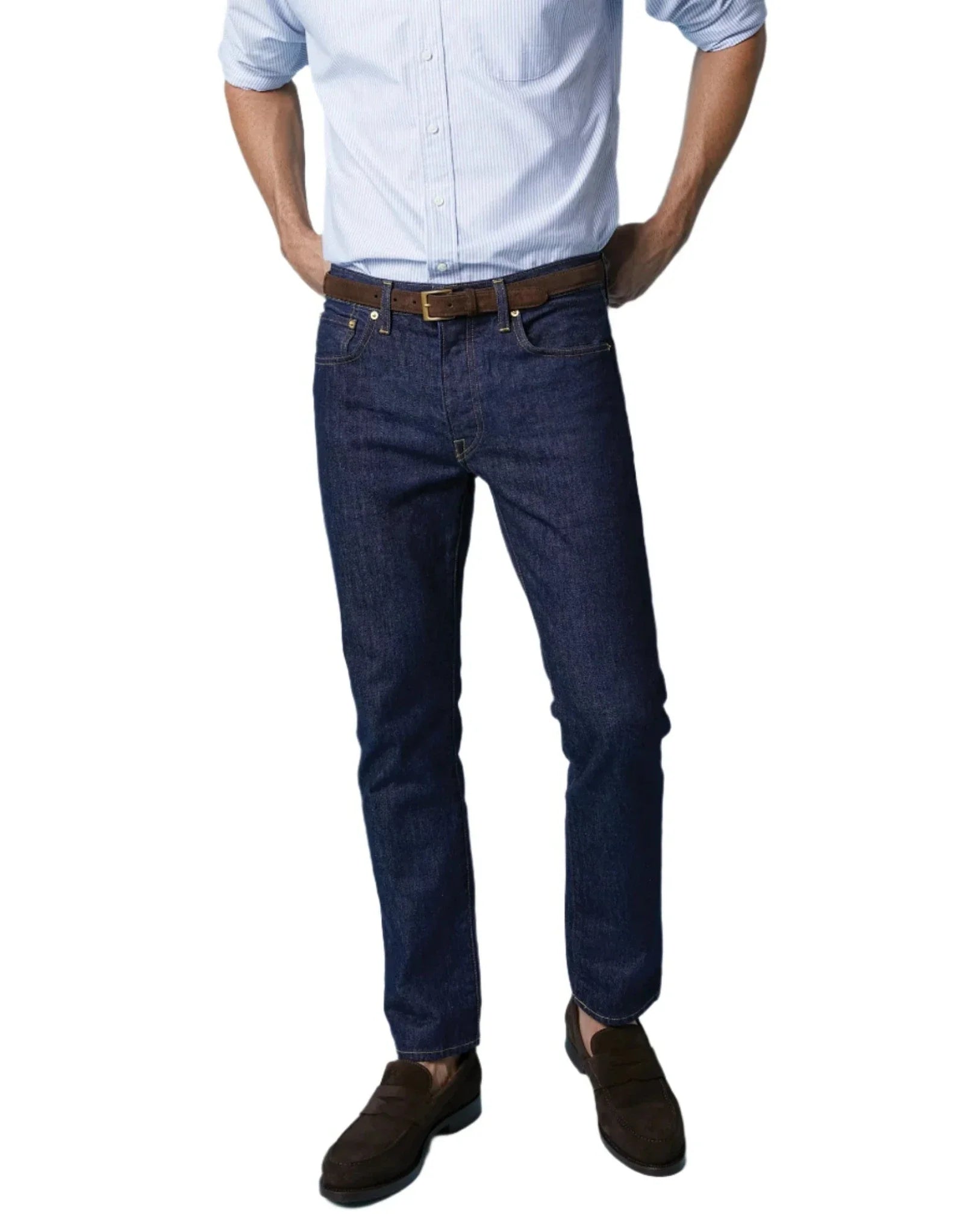 A person wearing a pair of Sid Mashburn Slim Straight Jeans in dark blue, crafted in the USA, paired with a light blue shirt, complemented by a brown belt and brown shoes, standing with hands in pockets.