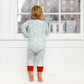 A child dressed in a Joy Street North Pole Christmas Baby Onesie stands on the windowsill, gazing out at the cityscape through a large window, dreaming of adventures at the North Pole.