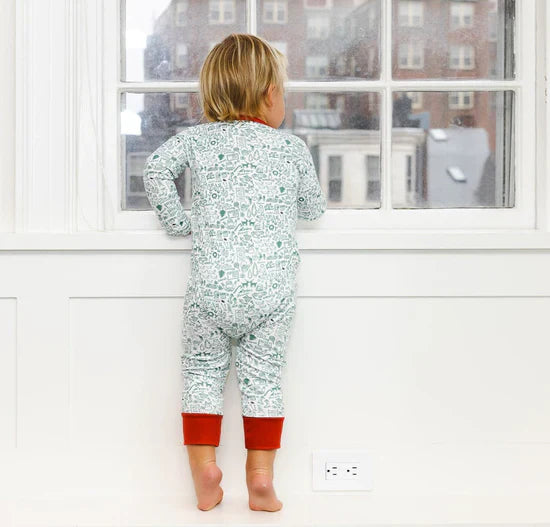 A child dressed in a Joy Street North Pole Christmas Baby Onesie stands on the windowsill, gazing out at the cityscape through a large window, dreaming of adventures at the North Pole.