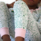 A close-up of someone wearing the Joy Street Kids North Pole Two Piece Henley Pajamas, featuring a white and green pattern with pink-striped cuffs, evoking the nostalgia of Christmas, while sitting with knees raised.