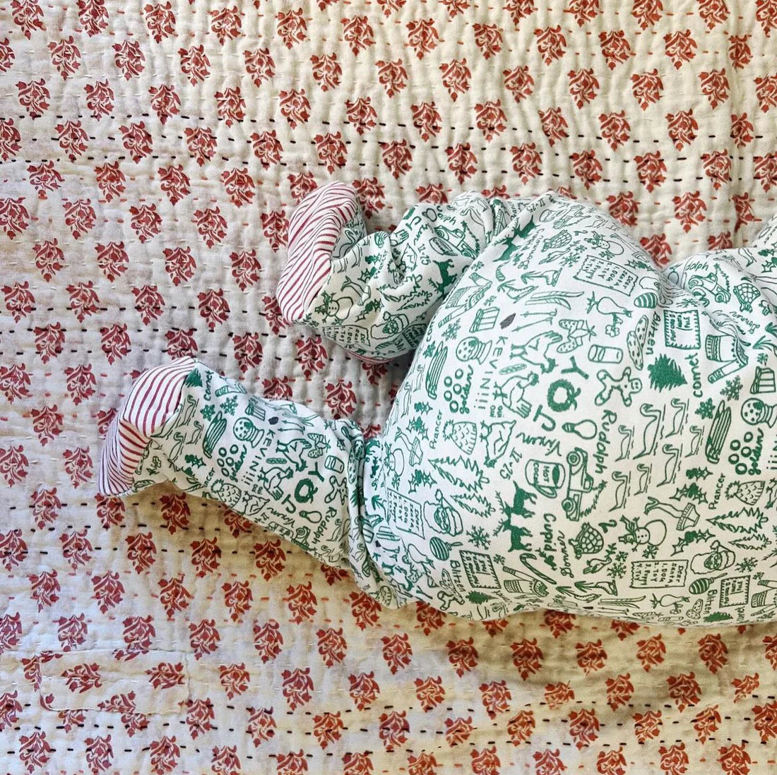 A baby wearing a Joy Street North Pole Christmas Baby Onesie cozily lies on a floral-patterned blanket.