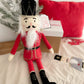 A plush MON AMI The Nutcracker Shelf Sitter - Red by Mon Ami in a red uniform sits on a white textured surface. In the background, you can see a decorated pine branch, a red cushion, and a wrapped gift box—all elements that make it the perfect holiday accent for your home decor.