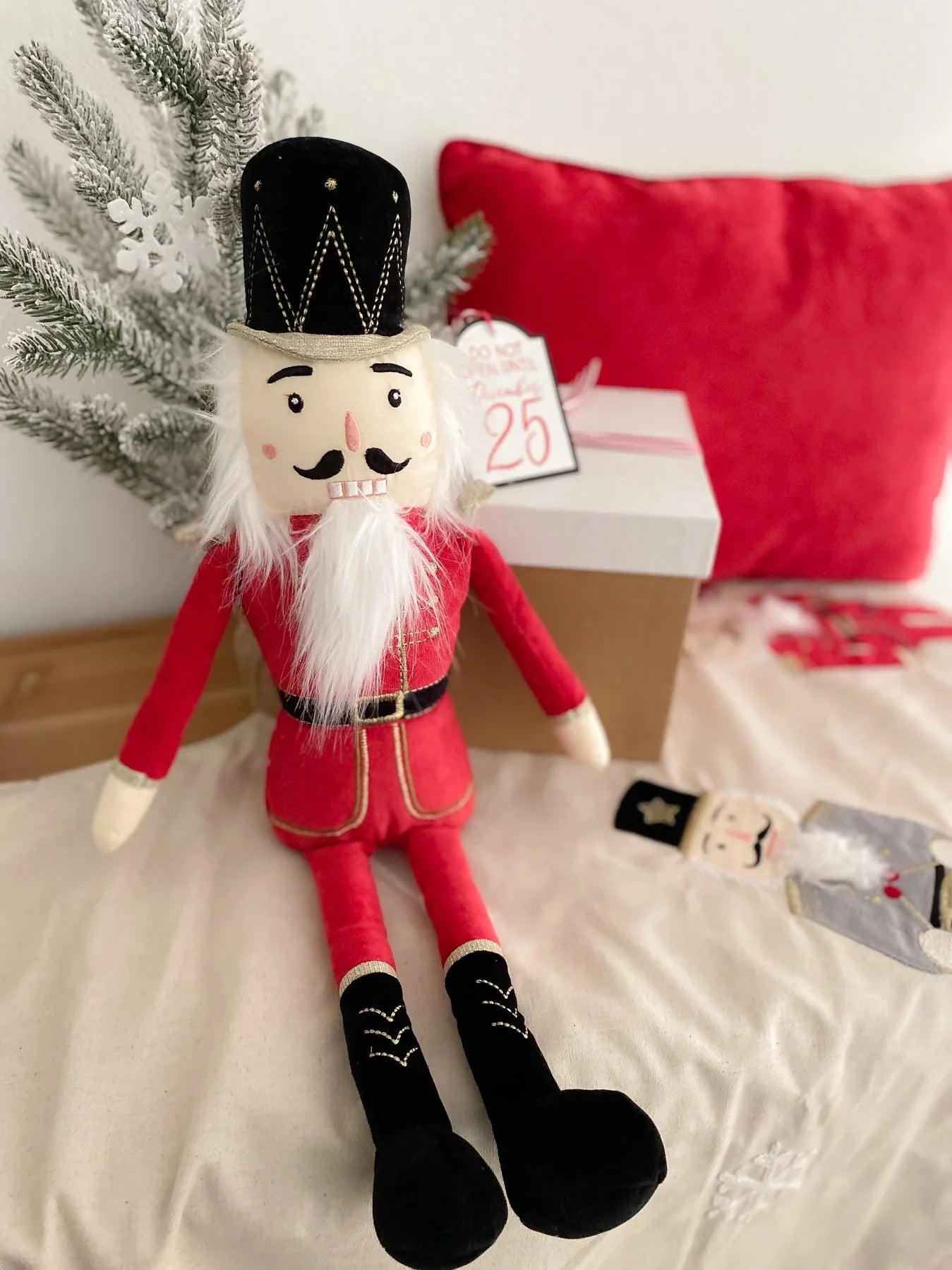 A plush MON AMI The Nutcracker Shelf Sitter - Red by Mon Ami in a red uniform sits on a white textured surface. In the background, you can see a decorated pine branch, a red cushion, and a wrapped gift box—all elements that make it the perfect holiday accent for your home decor.