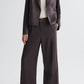 A person wears TWP’s Taite Shiny Viscose Wool Satin Pant in dark brown with high-rise wide-leg silhouette, paired with a white top and sandals, standing against a plain light background.