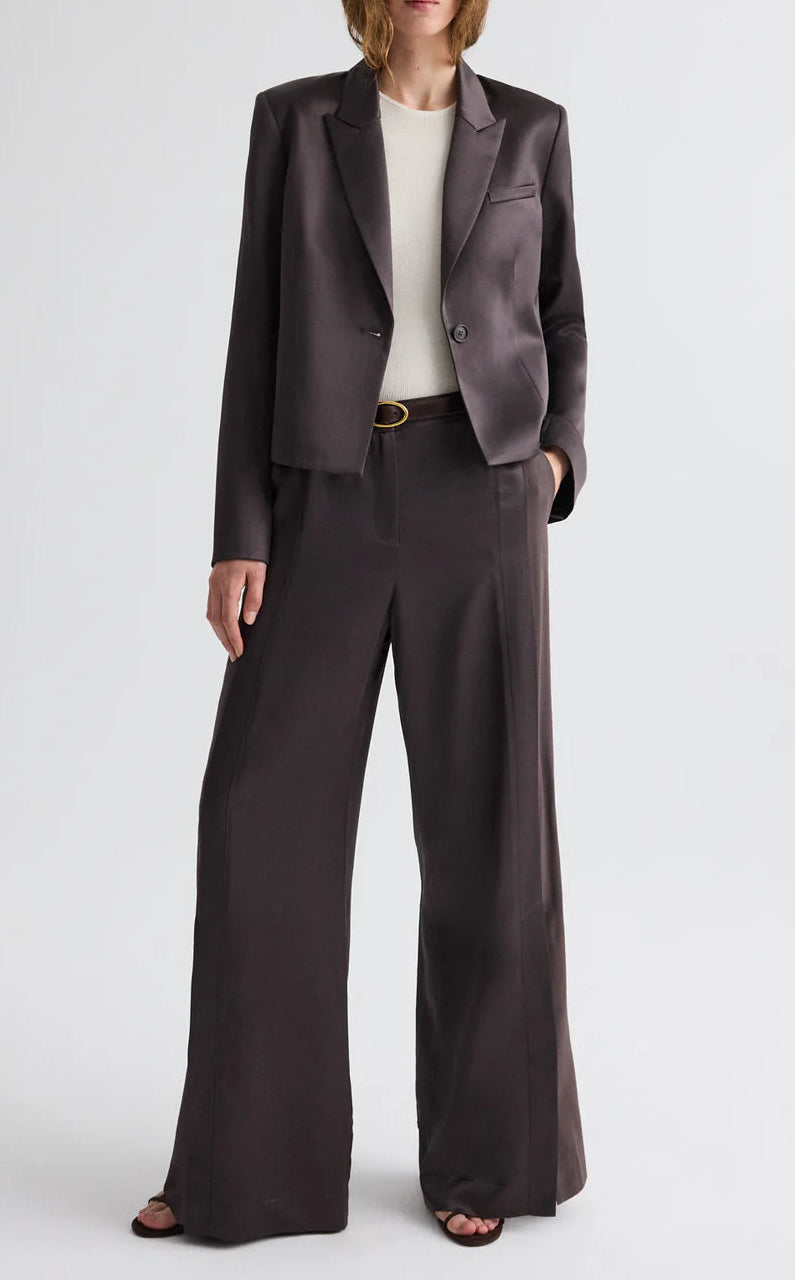 A person wearing the TWP Sawyer Shiny Viscose Wool Satin Blazer with matching wide-leg trousers, a white top, black sandals, and a belt stands against a plain background.