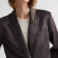 A model wears the TWP Sawyer Shiny Viscose Wool Satin Blazer, a dark, fitted cropped piece layered over a light ribbed shirt, shown from shoulders to mid-torso against a plain background.