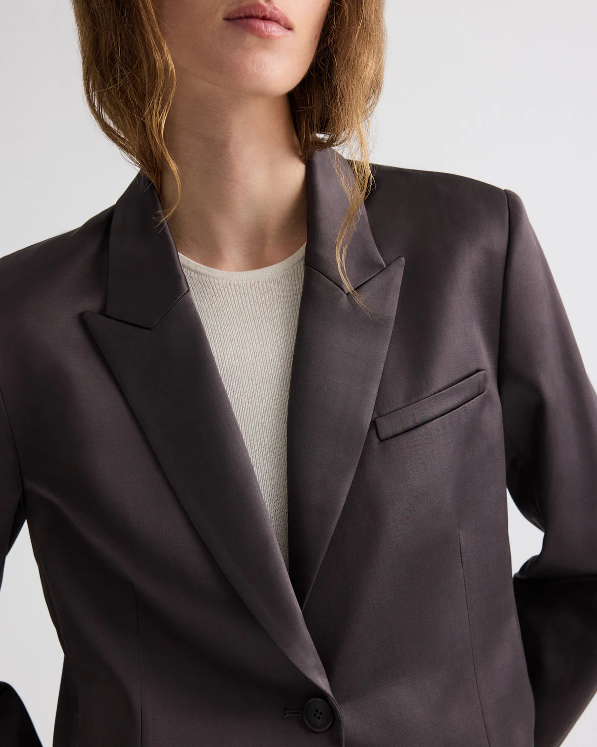 A model wears the TWP Sawyer Shiny Viscose Wool Satin Blazer, a dark, fitted cropped piece layered over a light ribbed shirt, shown from shoulders to mid-torso against a plain background.