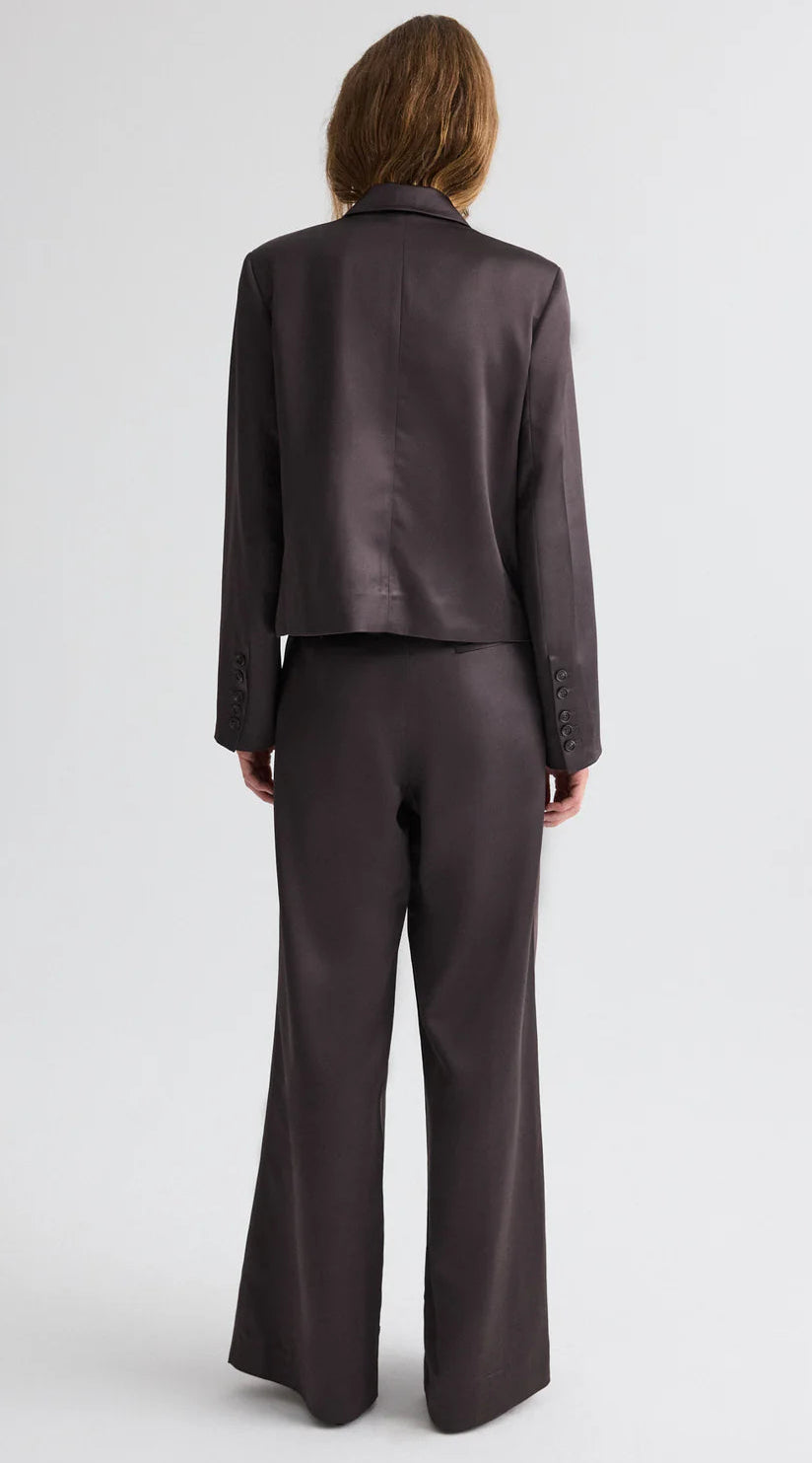 A person with shoulder-length hair is seen from the back, wearing the TWP Sawyer Shiny Viscose Wool Satin Blazer and matching wide-leg trousers against a plain light background.