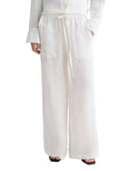 A person wears TWP’s Main St Washed Hemp Denim Pant—loose-fitting pull-ons with a drawstring waist, wide legs, and large front pockets; hands in pockets, white shirt and black sandals partly visible.