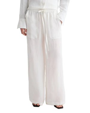 A person wears TWP’s Main St Washed Hemp Denim Pant—loose-fitting pull-ons with a drawstring waist, wide legs, and large front pockets; hands in pockets, white shirt and black sandals partly visible.