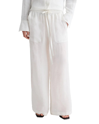 A person wears TWP’s Main St Washed Hemp Denim Pant—loose-fitting pull-ons with a drawstring waist, wide legs, and large front pockets; hands in pockets, white shirt and black sandals partly visible.