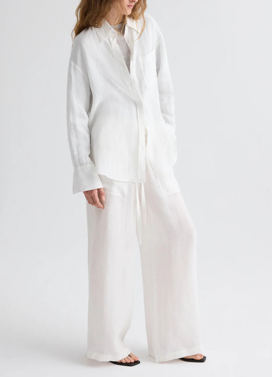 A person wearing a loose-fitting white long-sleeve shirt and TWP Main St Washed Hemp Denim Pant, styled with black sandals, stands against a plain light background.