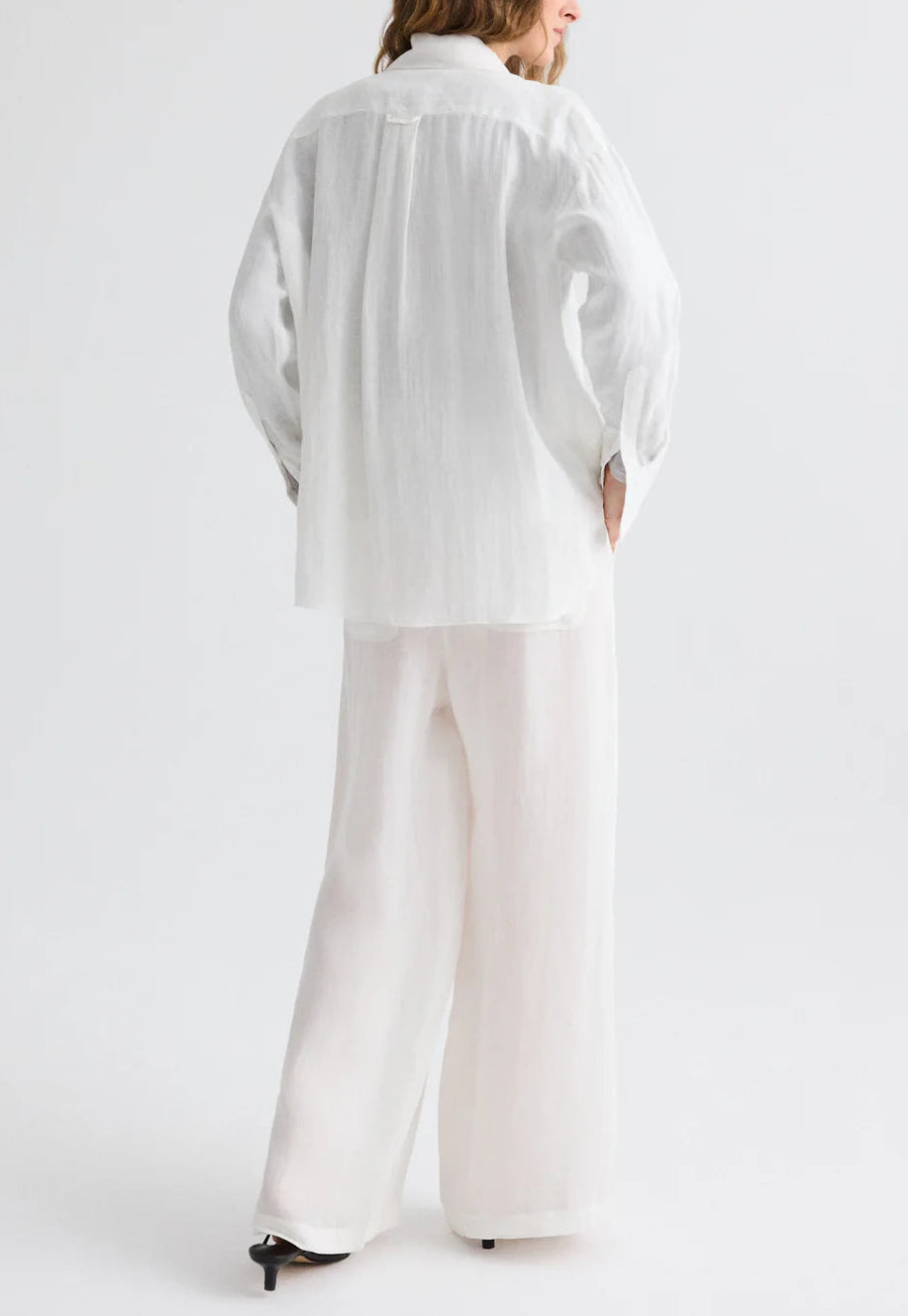 A person faces away, wearing a loose white long-sleeve shirt and TWP Main St Washed Hemp Denim Pants—lightweight, wide-leg pull-ons—paired with black-heeled shoes against a plain light background.