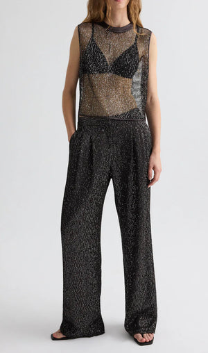 A person wears the TWP Sulli Sequin Netting Pant with a black bralette under a sleeveless, sheer metallic top and sandals.