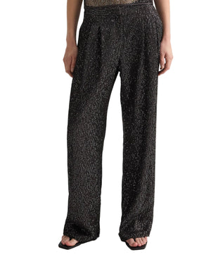 A person wearing TWP's Sulli Sequin Netting Pant—black, mid-rise wide-leg trousers with a subtle white dotted pattern—paired with open-toed black sandals.
