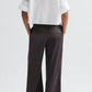 A person stands with hands in pockets, wearing a white cropped shirt and TWP's Taite Shiny Viscose Wool Satin Pant, high rise and wide leg, photographed from the back.