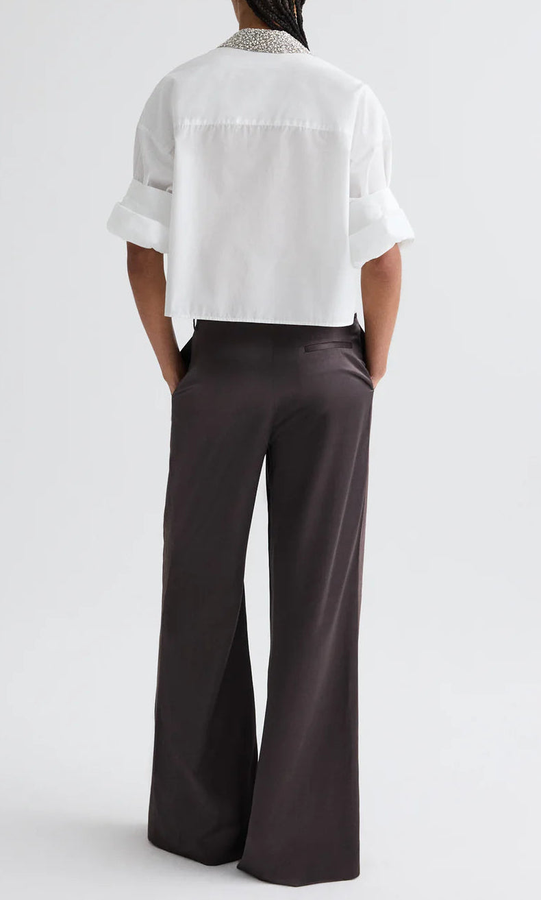 A person stands with hands in pockets, wearing a white cropped shirt and TWP's Taite Shiny Viscose Wool Satin Pant, high rise and wide leg, photographed from the back.