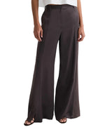 Model wears the TWP Taite Shiny Viscose Wool Satin Pant in dark brown, a high-rise, wide-leg style with front seams and side slits at the hem, paired with open-toe sandals.