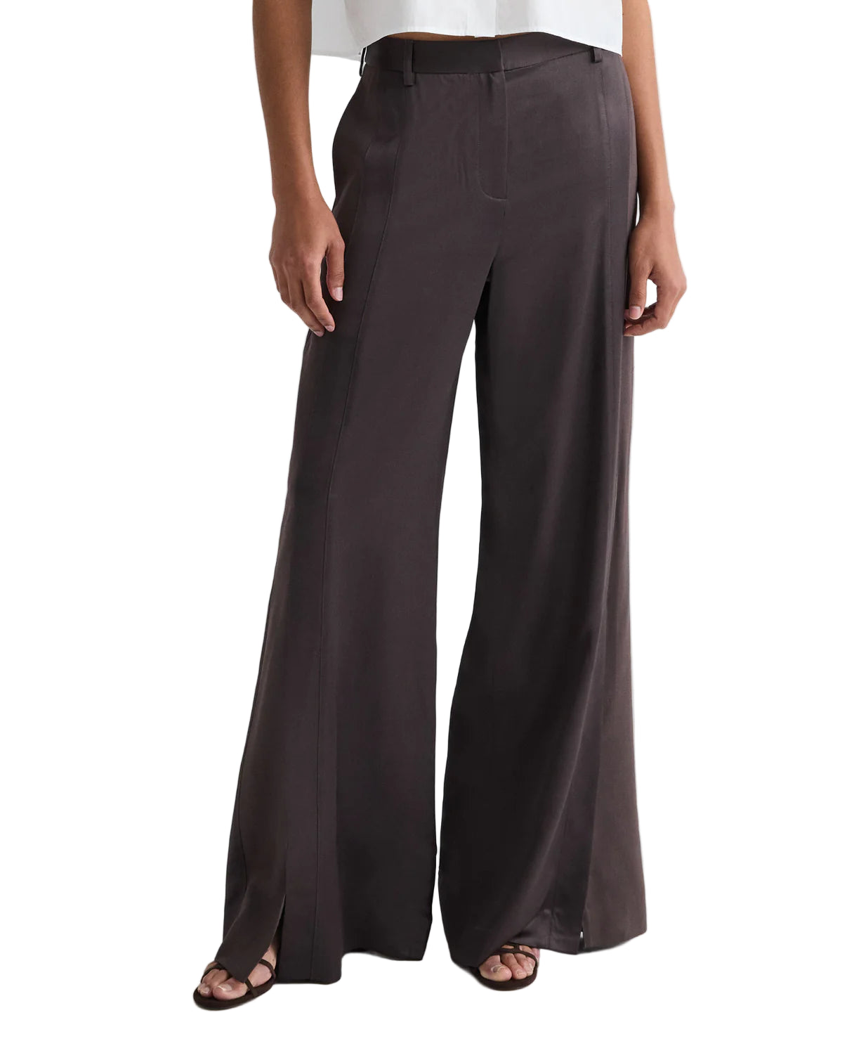 Model wears the TWP Taite Shiny Viscose Wool Satin Pant in dark brown, a high-rise, wide-leg style with front seams and side slits at the hem, paired with open-toe sandals.