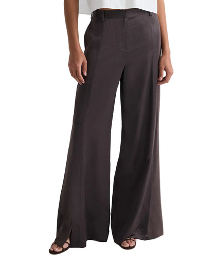 Model wears the TWP Taite Shiny Viscose Wool Satin Pant in dark brown, a high-rise, wide-leg style with front seams and side slits at the hem, paired with open-toe sandals.