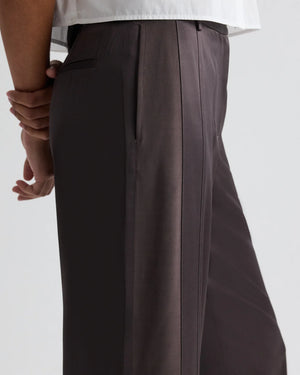 A person wearing TWP's Taite Shiny Viscose Wool Satin Pant in dark brown with high rise, wide leg, and side pocket, paired with a white shirt, shown from the waist down in a side view.