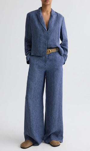 A person wearing TWP's Dylan Hemp Denim Pant in blue, styled with a beige belt and tan shoes, stands against a plain light background.