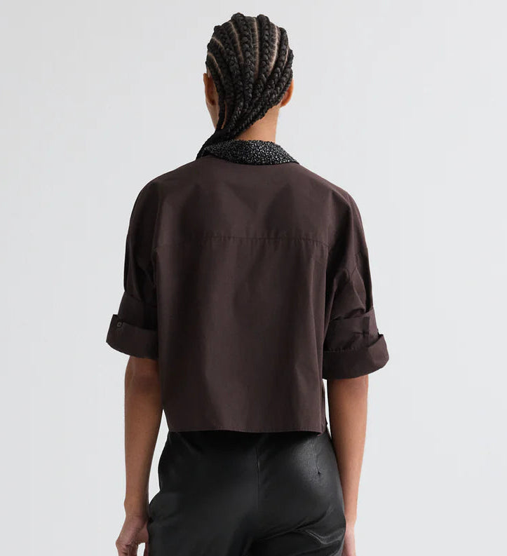 A person with braided hair, wearing the TWP Next Ex Crystal Collar Shirt in dark brown and black pants, stands with their back to a plain light background.