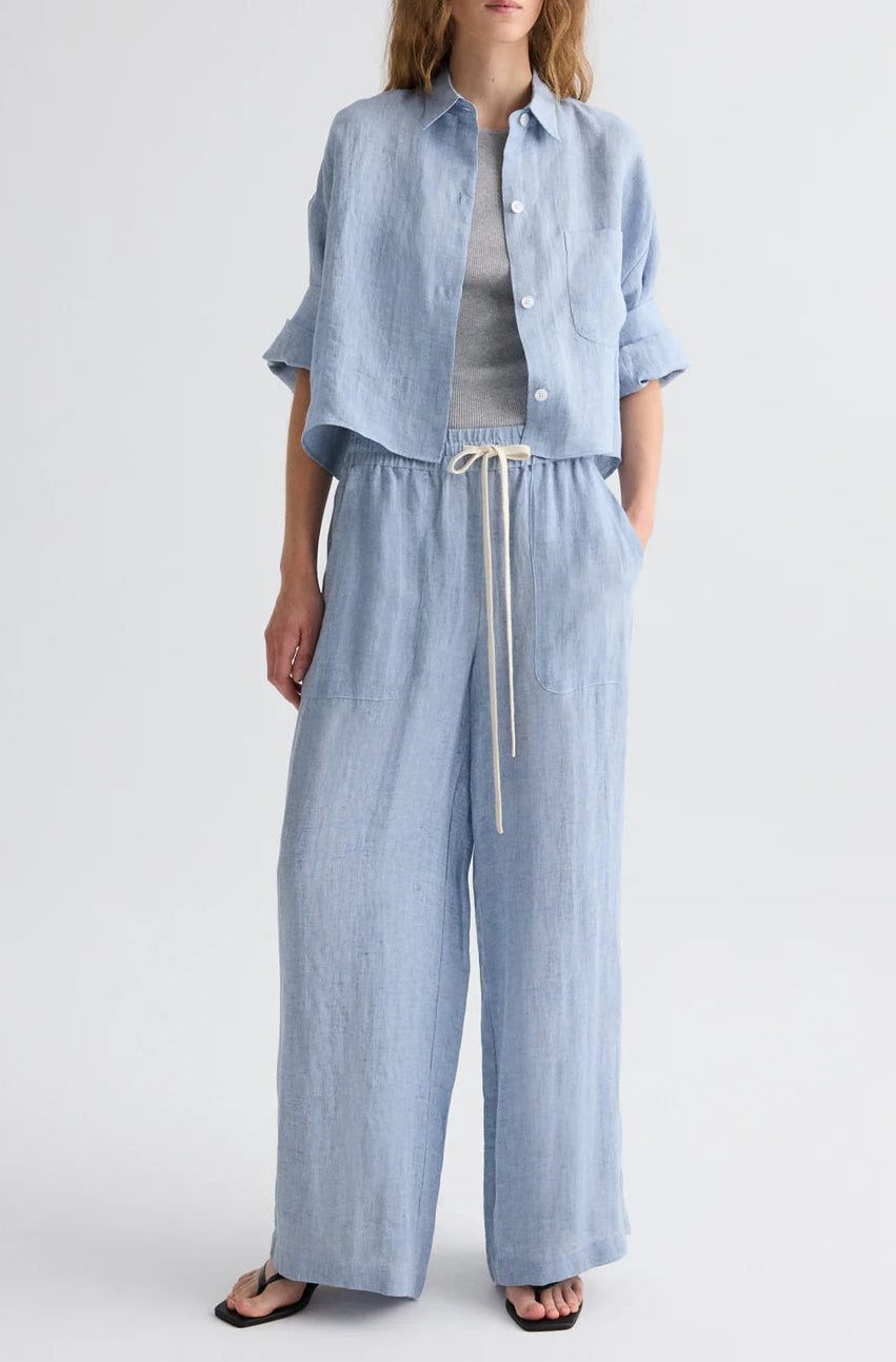 Someone wearing the TWP Next Ex Delave Hemp Shirt in light blue with a boxy fit, coordinated wide-leg drawstring pants, a grey tee, and black sandals stands against a plain background.