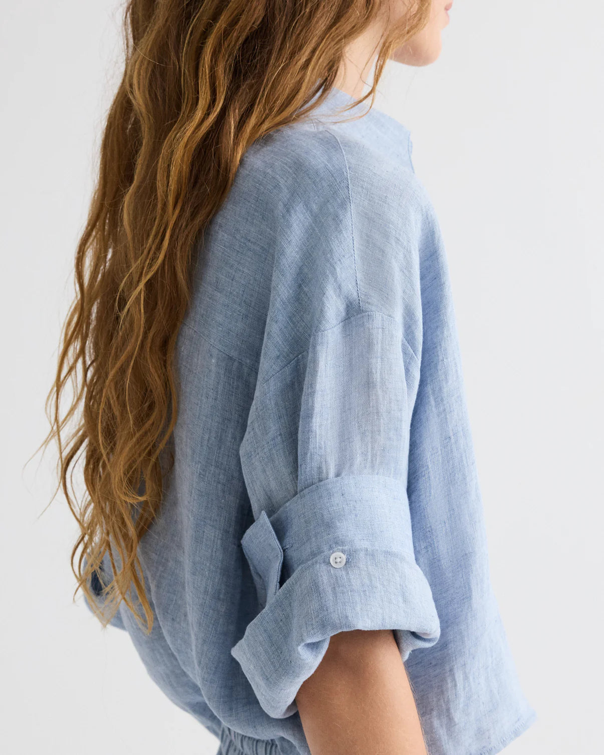 A person with long, wavy light brown hair models the TWP Next Ex Delave Hemp Shirt—a boxy, light blue linen top with rolled-up sleeves—shown in profile against a plain background.