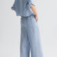 A person stands side-on to the camera, wearing the TWP Next Ex Delave Hemp Shirt—a cropped, light blue linen top with a boxy fit—paired with matching wide-leg pants and black heeled sandals.