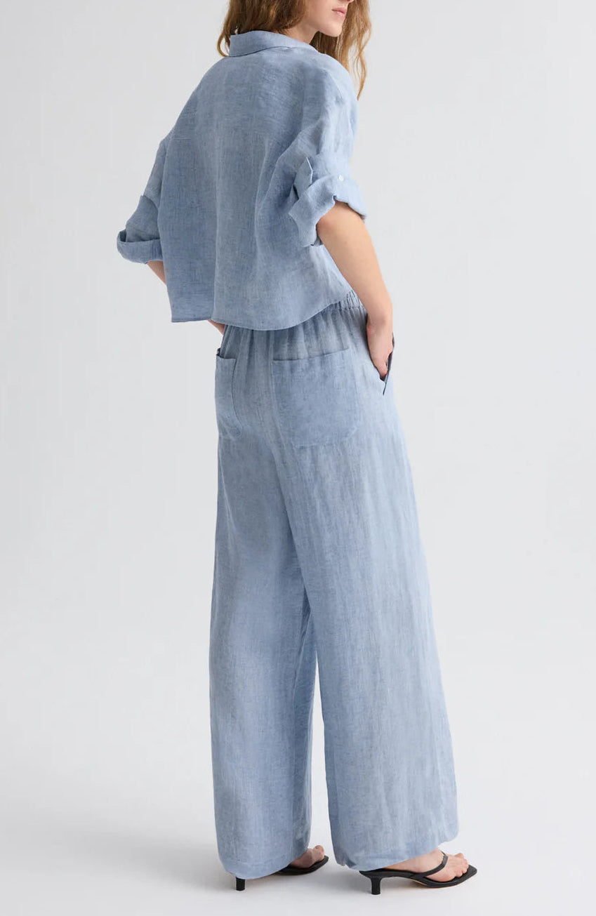 A person stands side-on to the camera, wearing the TWP Next Ex Delave Hemp Shirt—a cropped, light blue linen top with a boxy fit—paired with matching wide-leg pants and black heeled sandals.