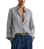A person wears the TWP Next Morning Clear Sequin Stripe Shirt, crafted from Italian cotton shirting in blue and white stripes, partially unbuttoned, styled with blue pants and a brown belt. Hands are in their pockets.