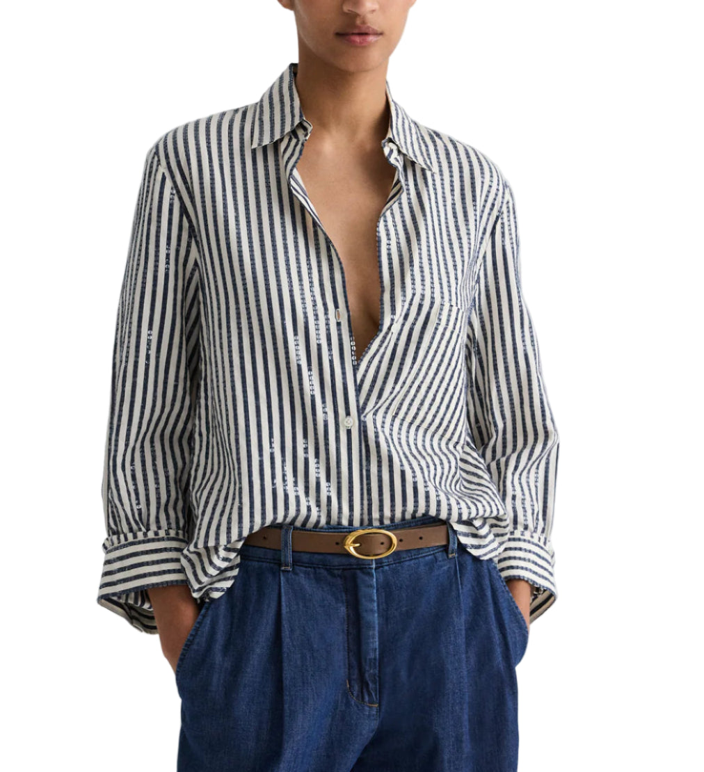 A person wears the TWP Next Morning Clear Sequin Stripe Shirt, crafted from Italian cotton shirting in blue and white stripes, partially unbuttoned, styled with blue pants and a brown belt. Hands are in their pockets.