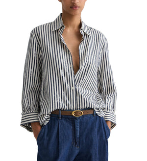 A person wears the TWP Next Morning Clear Sequin Stripe Shirt, crafted from Italian cotton shirting in blue and white stripes, partially unbuttoned, styled with blue pants and a brown belt. Hands are in their pockets.