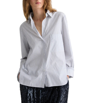 A person wears the TWP Next Morning Micro Hairline Stripe Shirt—an oversized, light blue and white striped button down—with hands in pockets, paired with a black sequin skirt. The background is white.