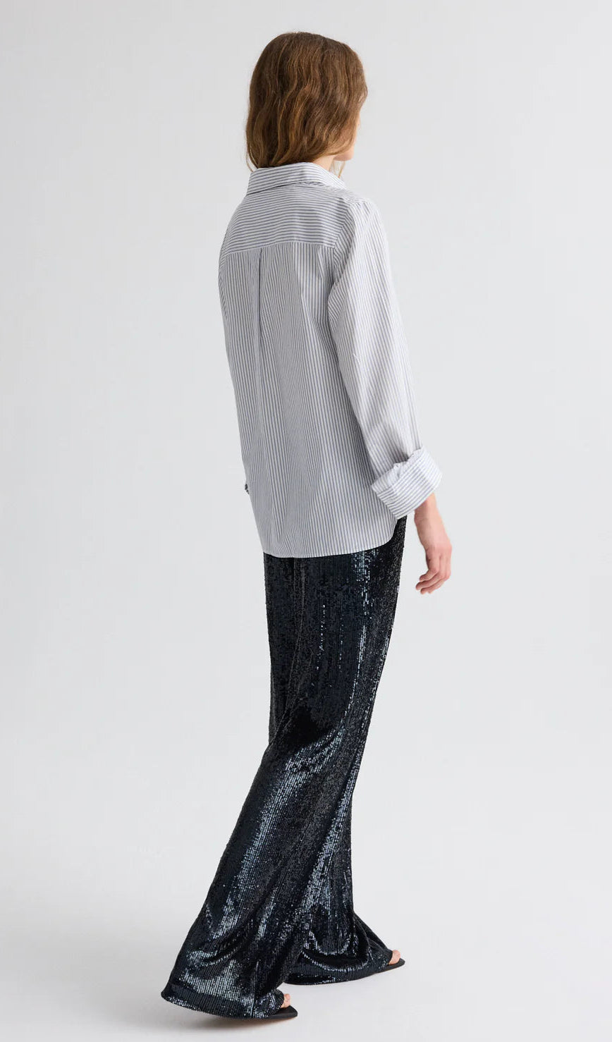 A person faces away in the TWP Next Morning Micro Hairline Stripe Shirt—an oversized button-down with rolled sleeves—and black sequin flared pants, set against a plain background.