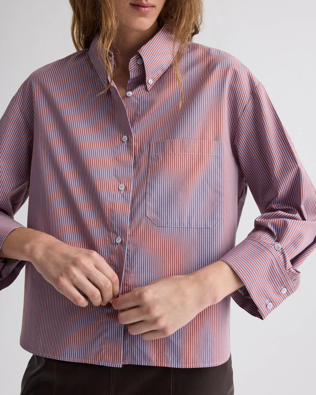A person stands buttoning the TWP J.D. Framed Micro Striped Shirt, a boxy-fit long sleeve shirt in red and white Italian cotton shirting.