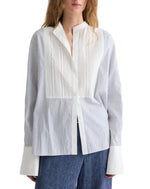 A woman wearing TWP’s The Night Before Cotton Stripes Shirt, a white and light blue long-sleeve button-up with a band collar, wide cuffs, pintuck details, paired with dark blue pants.