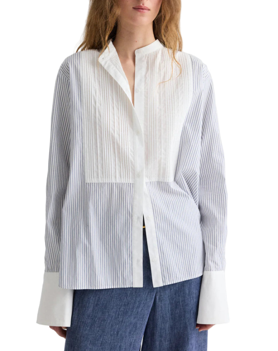 A woman wearing TWP’s The Night Before Cotton Stripes Shirt, a white and light blue long-sleeve button-up with a band collar, wide cuffs, pintuck details, paired with dark blue pants.