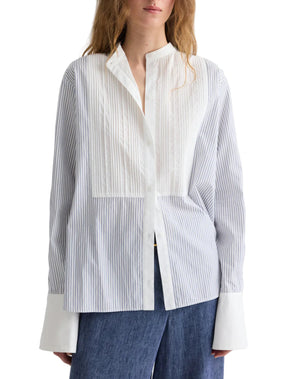 A woman wearing TWP’s The Night Before Cotton Stripes Shirt, a white and light blue long-sleeve button-up with a band collar, wide cuffs, pintuck details, paired with dark blue pants.