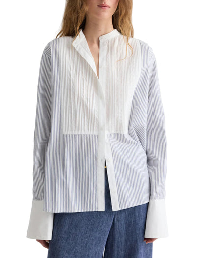 A woman wearing TWP’s The Night Before Cotton Stripes Shirt, a white and light blue long-sleeve button-up with a band collar, wide cuffs, pintuck details, paired with dark blue pants.