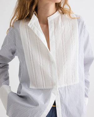A woman wears TWP's The Night Before Cotton Stripes Shirt, featuring a pleated white bib front, pintuck details, a mandarin collar, and long sleeves; her hand rests in her pocket.
