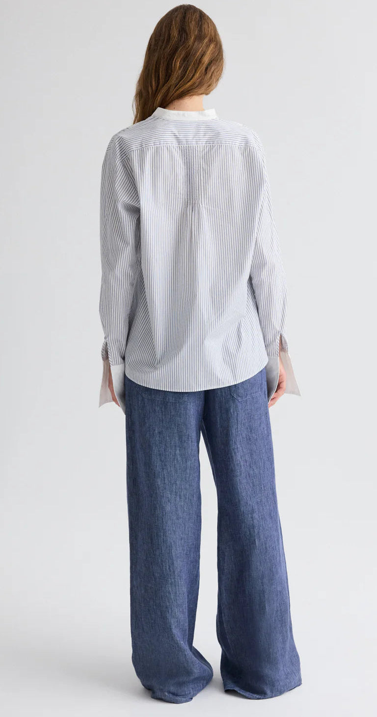 A person stands with their back to the camera, wearing TWP's The Night Before Cotton Stripes Shirt—a light blue striped long-sleeve button-up—paired with loose-fitting blue jeans against a plain background.