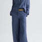 A person wearing the Lucy Hemp Denim Top by TWP, styled with wide-leg trousers, a beige belt, and brown shoes, stands against a plain light background.