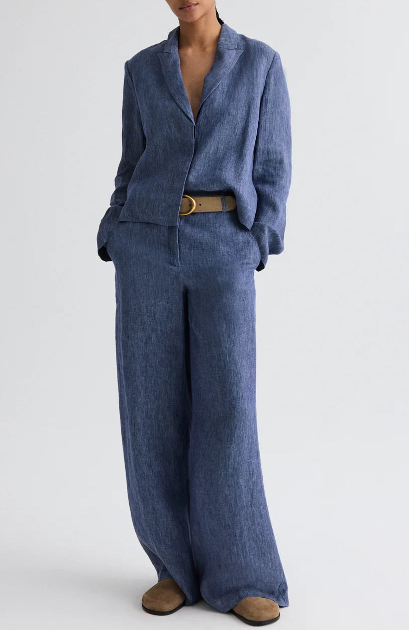 A person wearing the Lucy Hemp Denim Top by TWP, styled with wide-leg trousers, a beige belt, and brown shoes, stands against a plain light background.