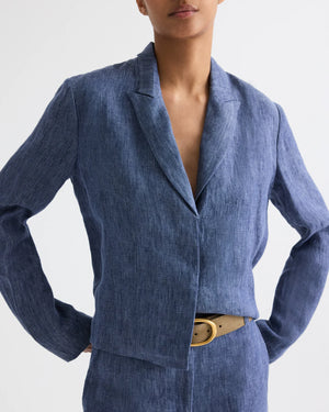 A person wearing the TWP Lucy Hemp Denim Top in blue, styled with matching pants and a beige belt, stands hands on hips against a plain background.